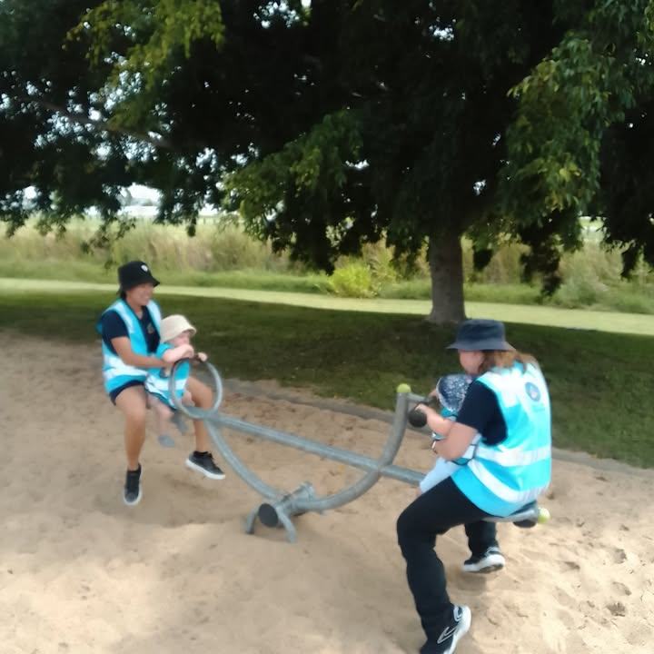 Two educators in blue excursion vests riding a see-saw at the park with toddlers held safely in front of them