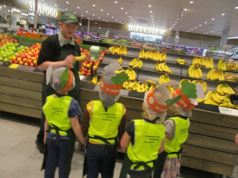 Children in hair nets, aprons and high-visibility vests standing with a greengrocer beside the banana display in a supermarket, photographed from behind