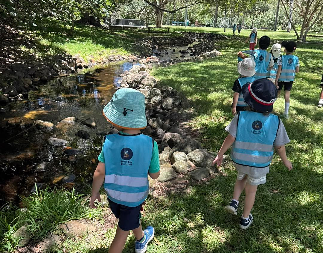 Children in high-visibility vests and sun hats walking beside a rock-lined creek in a shady park, photographed from behind