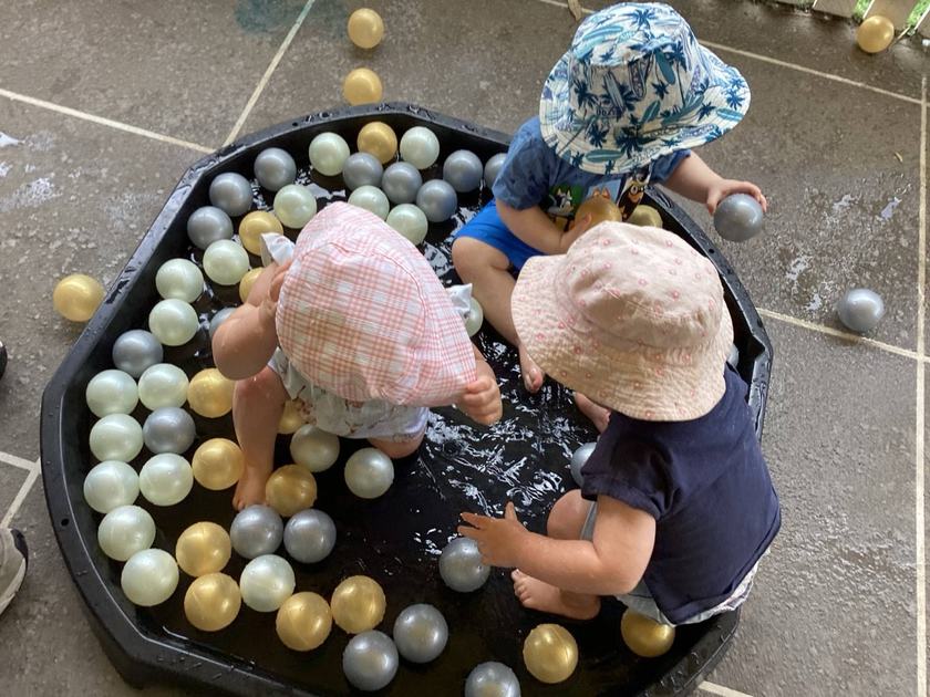 Three toddlers in wide-brim sun hats crouched around a black tuff tray of shallow water filled with gold, silver and pearl balls, on wet paving at one of our centres outdoor area