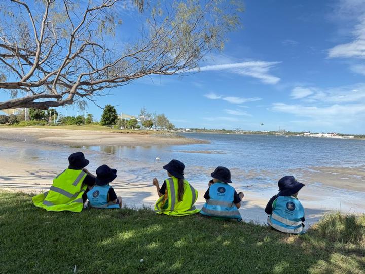 Five children in sun hats and high-vis safety vests sitting on grass in the shade of a low tree at the water's edge, photographed from behind the group