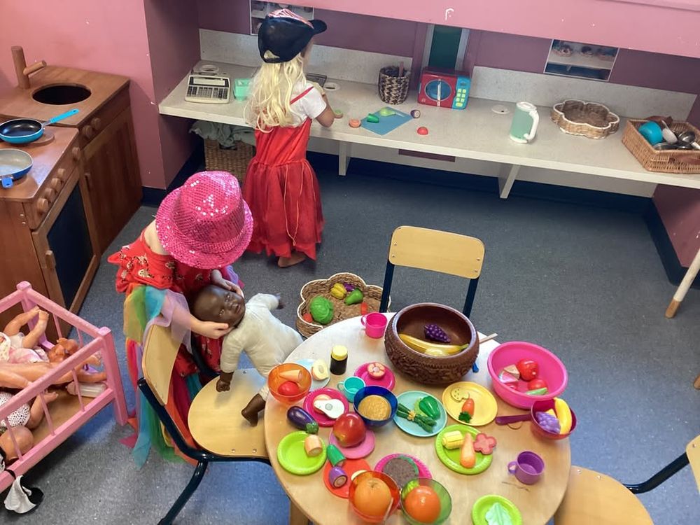 Two children in dress-ups playing in the home corner, one at a table set with play food and plates and the other at a bench with a toy cash register, photographed from behind