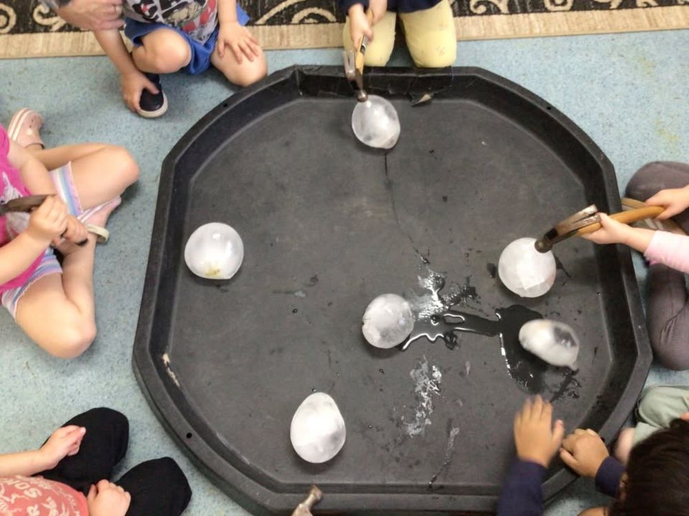 Children sitting around a large black tray, using small wooden hammers to crack open balls of ice, photographed from directly above so only their hands are in frame