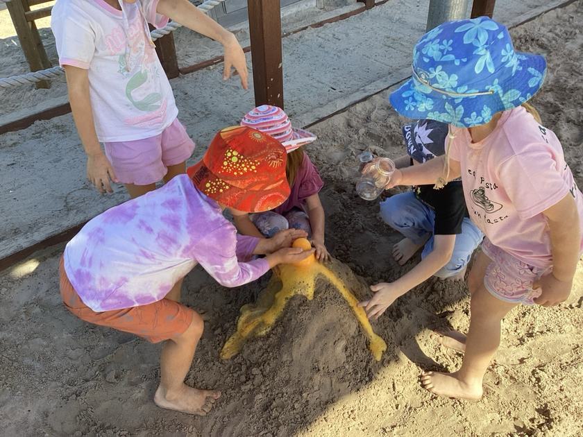 Four children in wide-brim sun hats crouched around a mound of damp sand in a sandpit, one tipping sand from a container onto the pile